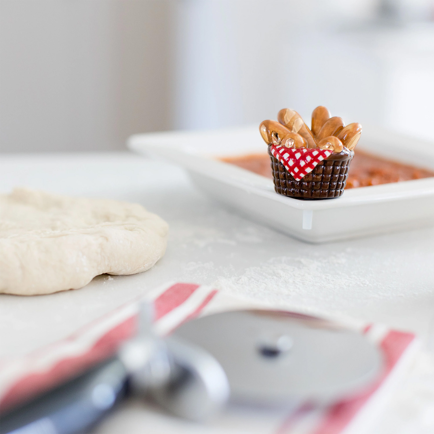 Nora Fleming mini You Knead it shaped like a basket of bread, attached to a serving bowl base filled with pizza sauce. There is pizza dough and a pizza cutter in the foreground.