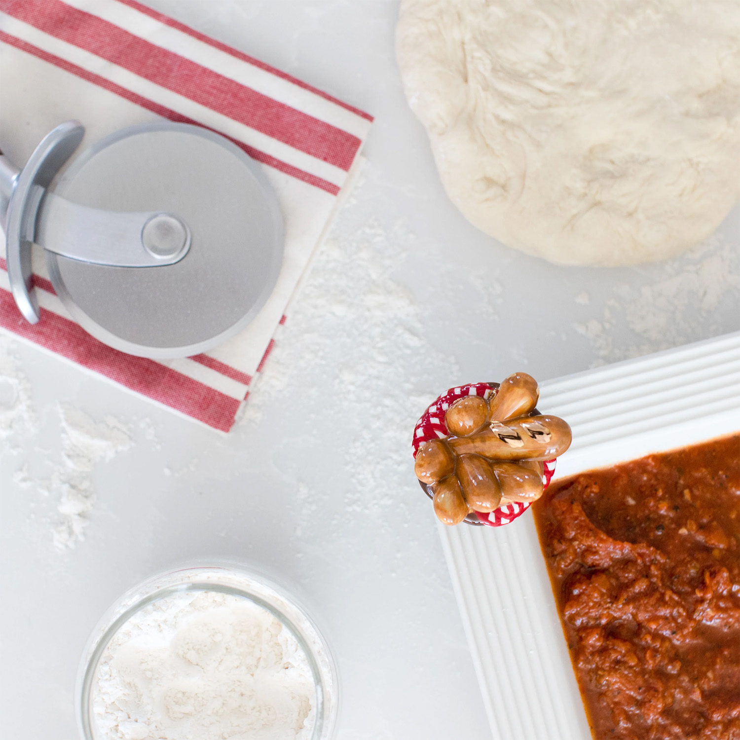 top view of a scene with pizza dough, a pizza cutter, and a Nora Fleming mini You Knead it shaped like a basket of bread attached to a bowl base filled with pizza sauce